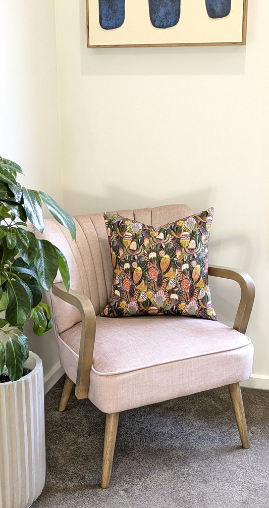A light pink armchair with wooden legs sits beside a green potted plant. On the chair rests the Emotive Collection’s Bush Banksia Cushion Cover. Above, a framed abstract painting is displayed.