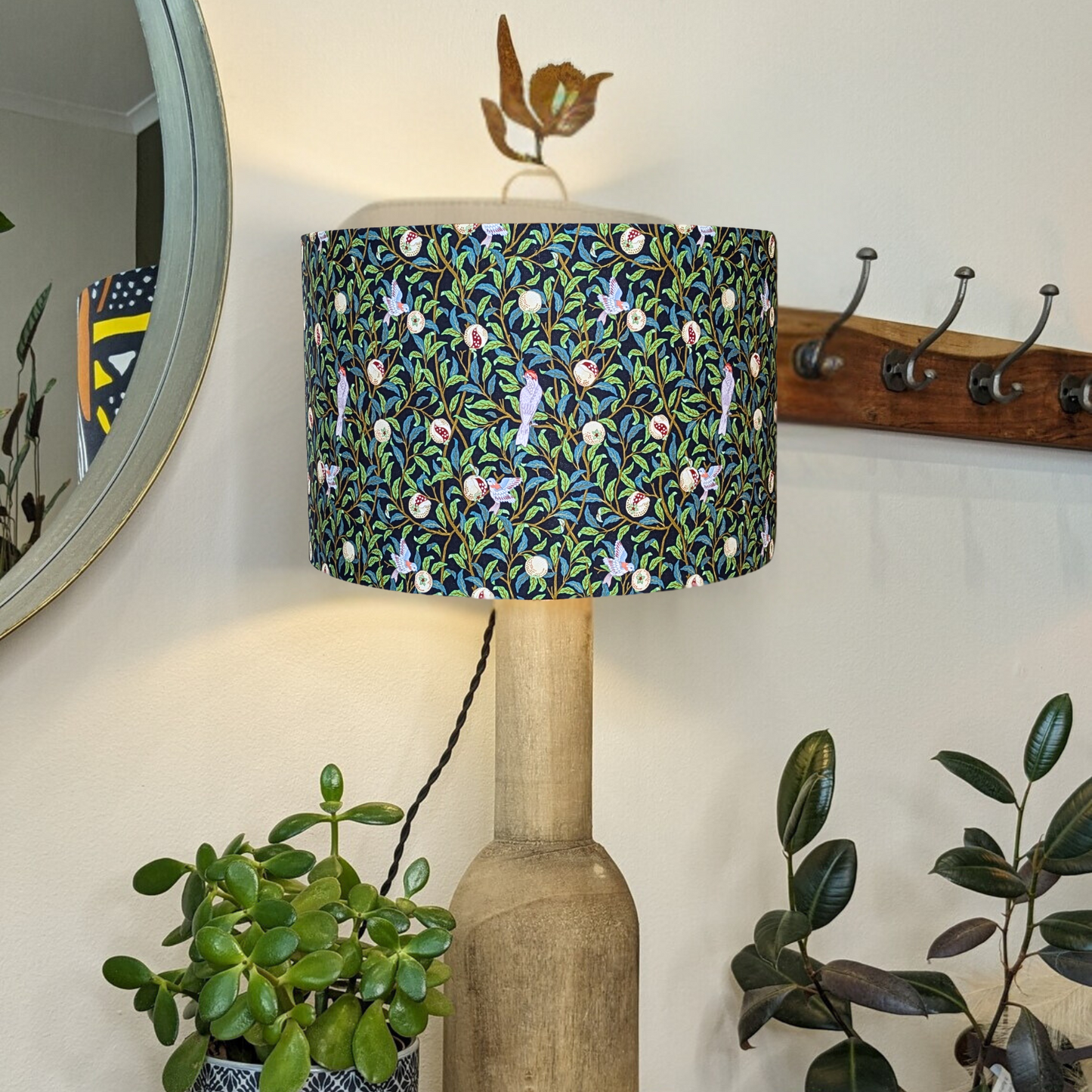 Handmade Bird & Pomegranate Black William Morris lampshade by Emotive Collection, shown on a wooden lamp base and the light is switched on, featuring a striking black background with vibrant bird and pomegranate motifs in the classic Morris style. Crafted