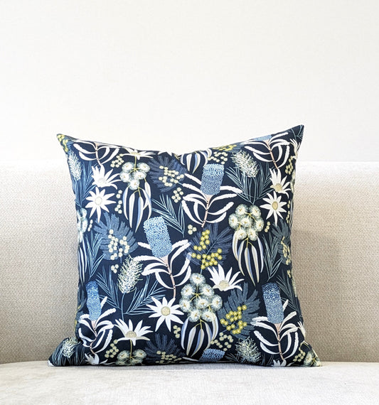 The Emotive Collection Blue Native Cushion Cover, with a dark blue botanical design inspired by Jocelyn Proust in white, yellow, and green, is displayed on a light beige sofa against a plain wall.