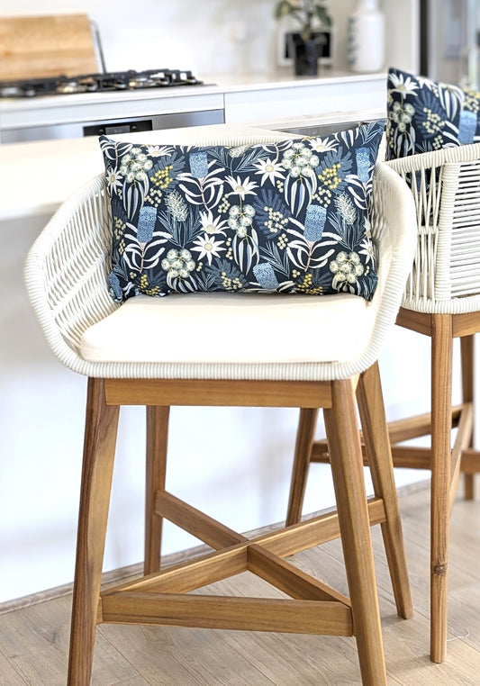 A wooden bar stool with a white woven seat is topped with the Emotive Collection’s Blue Native Cushion Cover, featuring a blue and white floral design, shown in a bright kitchen setting.