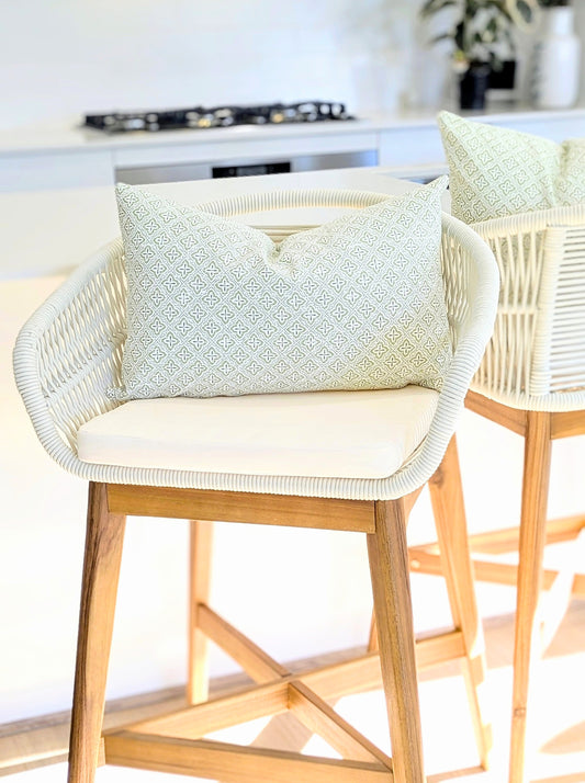 A bright kitchen features two white wicker barstools with wooden bases, each topped with an Emotive Collection Tiles Cocoon Cushion Cover in a light pattern. A plant and stovetop are softly visible in the background.