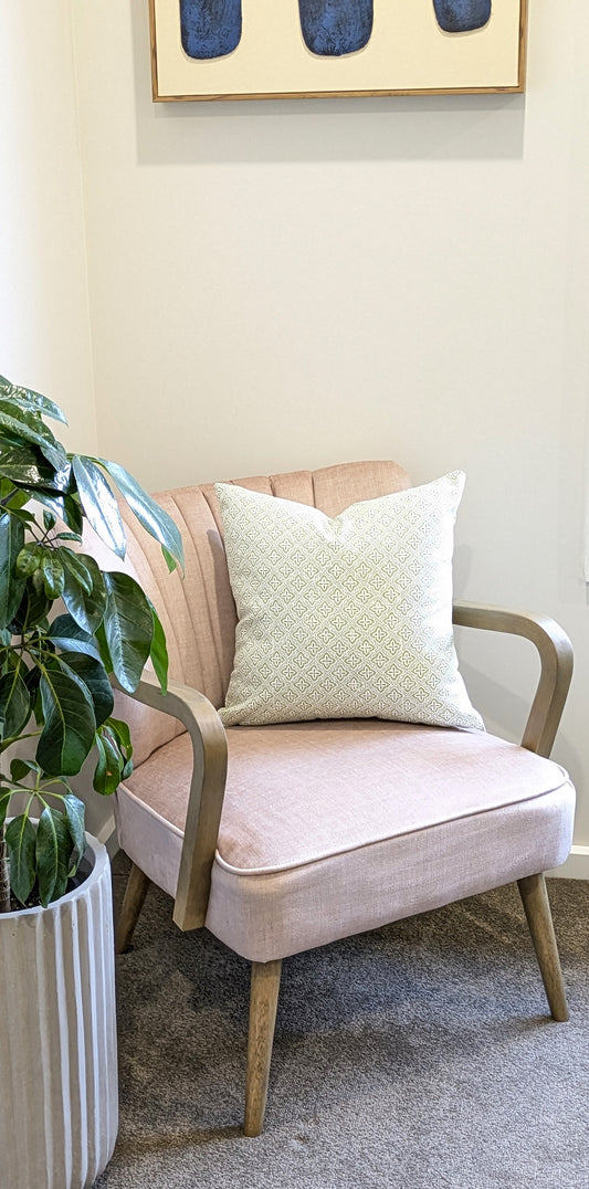 A light pink armchair with wooden arms and legs sits in a corner. The Tiles Cocoon Cushion Cover by Emotive Collection rests on the chair. A tall green potted plant from Emotive Collection is placed nearby on the left.