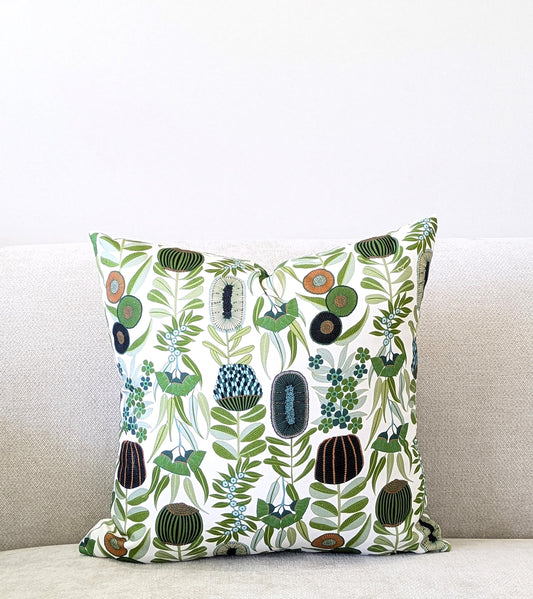 The Emotive Collection Green Australiana Cushion Cover, featuring a vibrant botanical pattern of green leaves and stylized flowers, is displayed on a light sofa against a plain white wall.