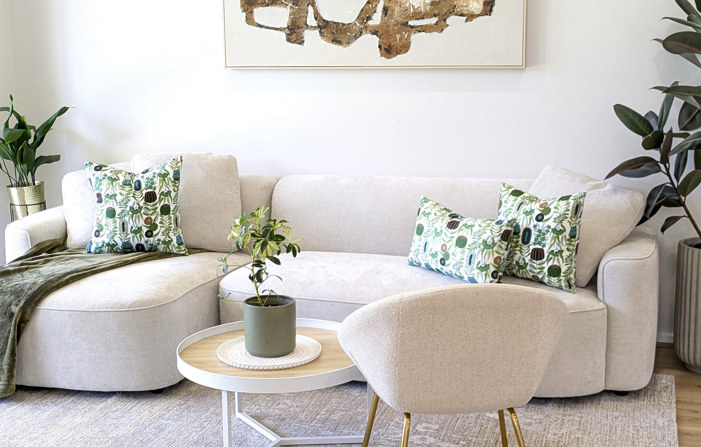 A modern living room features a light beige sectional sofa with Emotive Collection’s Green Australiana Cushion Covers, a white round coffee table with a potted plant, lush greenery, and abstract art on crisp white walls.