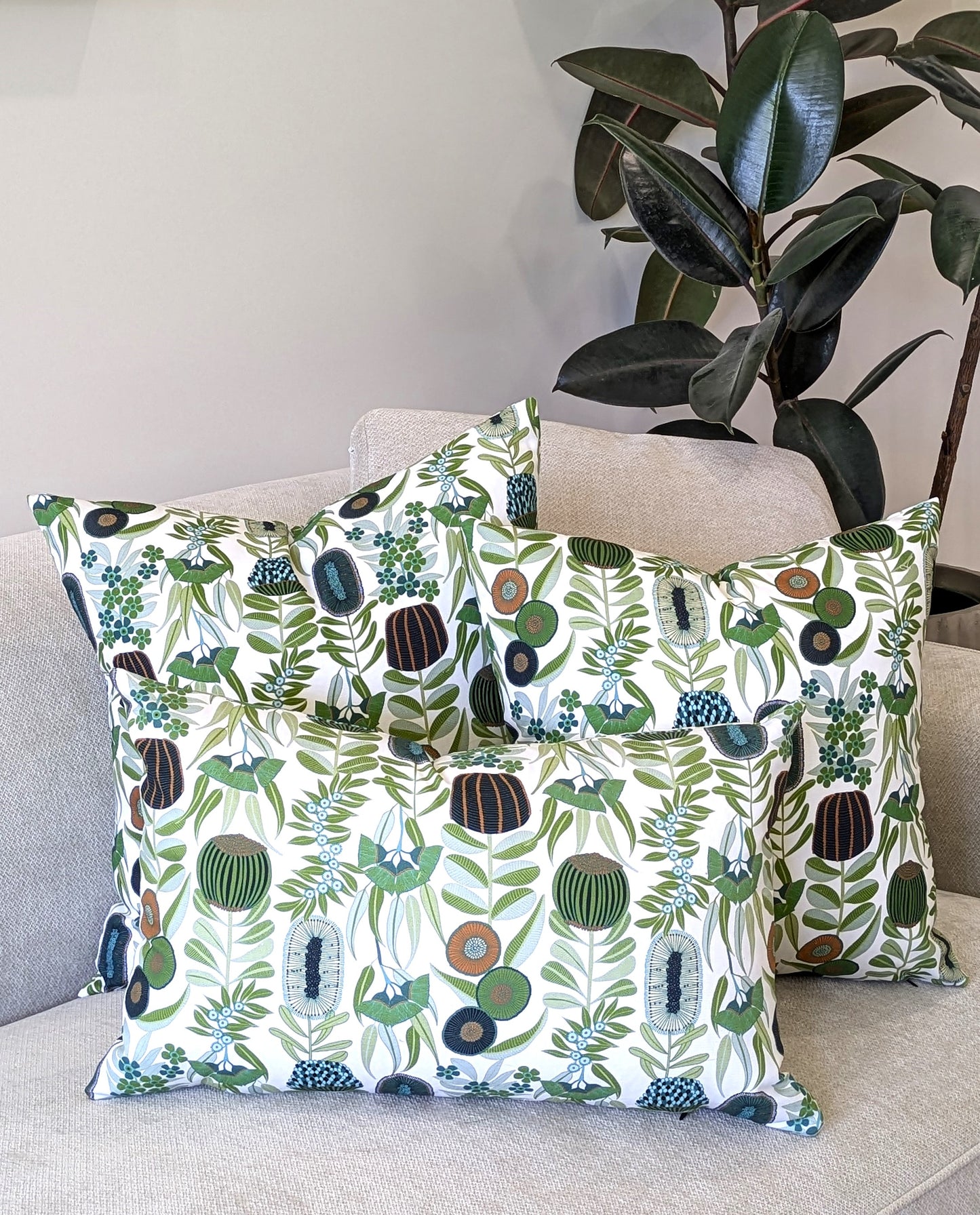 Three Emotive Collection Green Australiana Cushion Covers decorate a light beige sofa, featuring green, brown, and white botanical patterns inspired by Jocelyn Proust, with a leafy indoor plant in the background.
