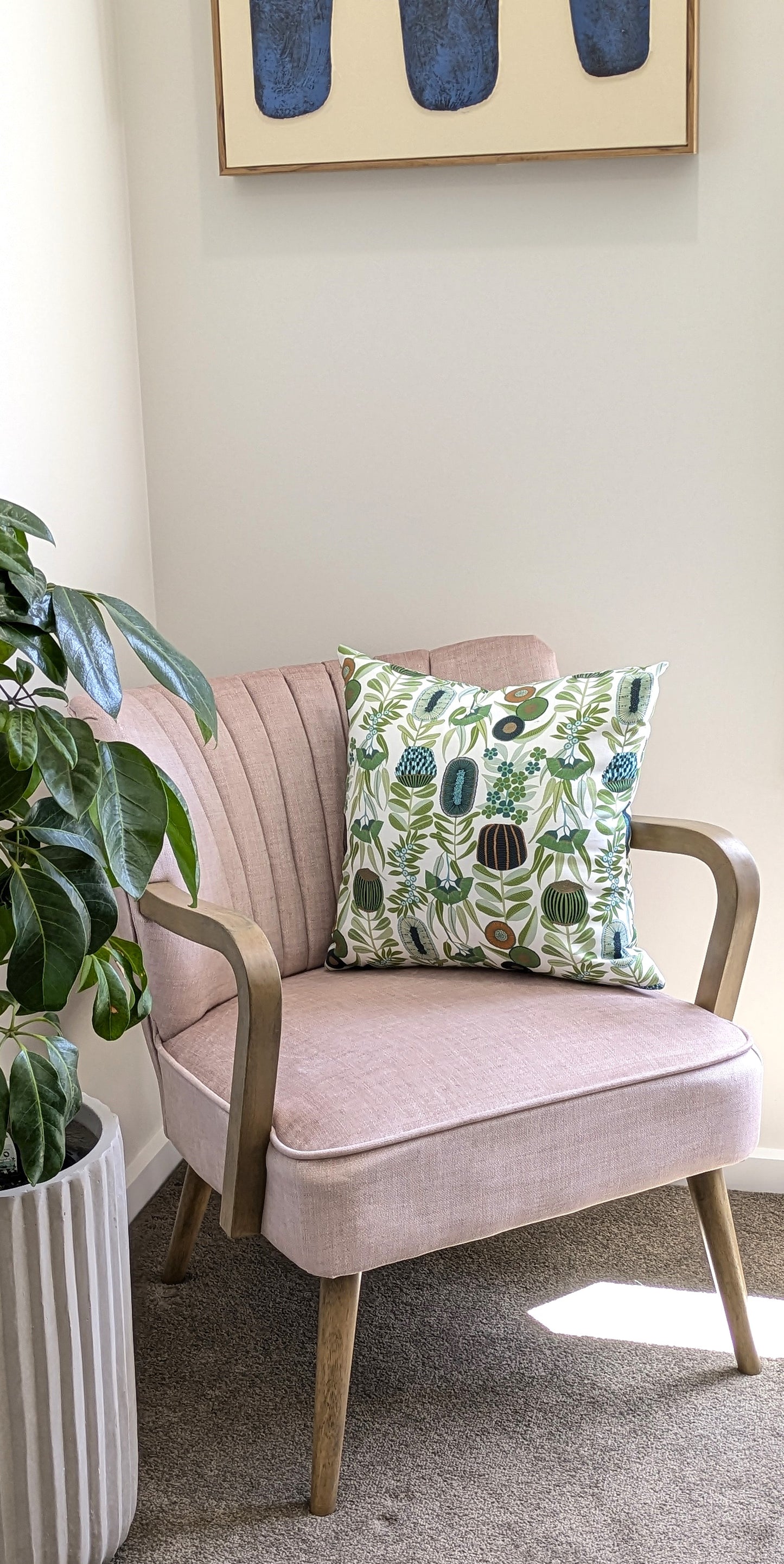 A light pink armchair with wooden legs and arms sits in a corner. It features the Green Australiana Cushion Cover from Emotive Collection, decorated with green and blue by Jocelyn Proust. A leafy plant and abstract art complete the scene.