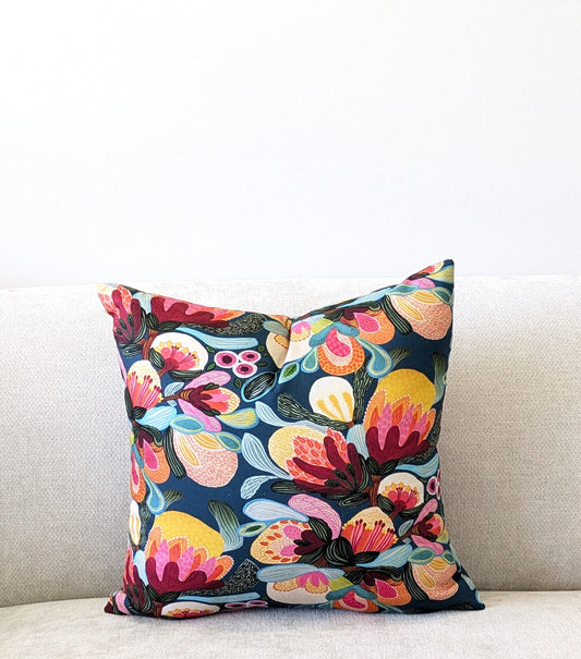 The Emotive Collections Midnight Bush Cushion Cover, with its vibrant floral pattern in pink, red, yellow, blue, and green, by Kirsten Katz Australia, brightens a light beige sofa set against a plain white wall.