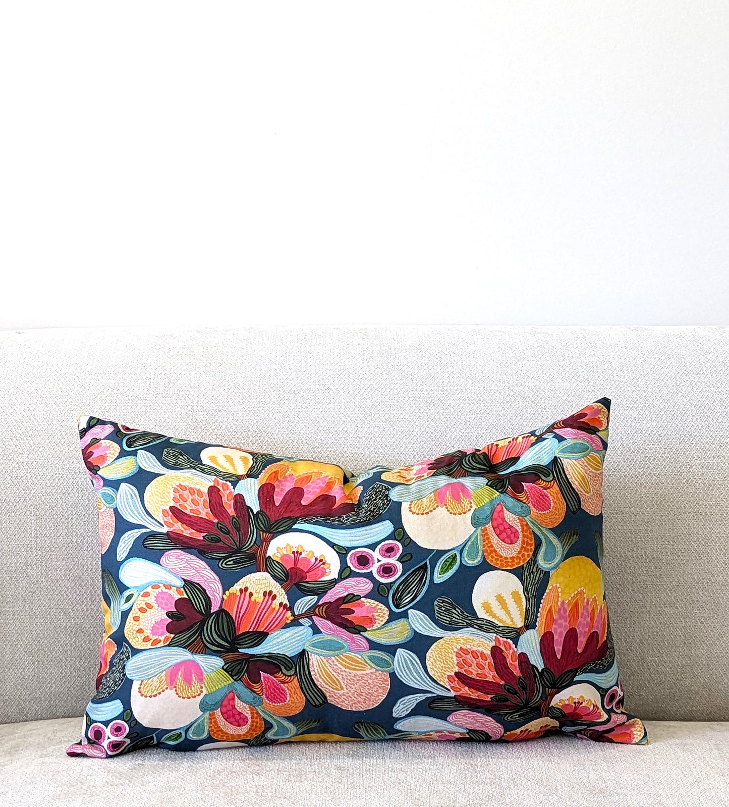 The Emotive Collection’s Midnight Bush Cushion Cover, featuring a vibrant floral design, is displayed on a light gray upholstered sofa against a plain white wall.