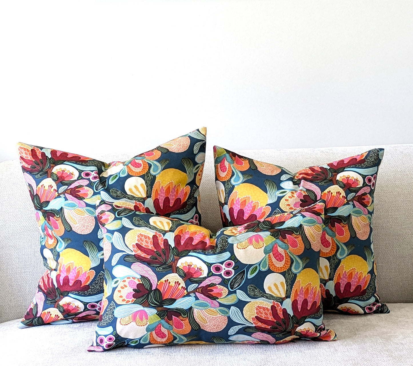 Four decorative pillows with vibrant multicolored floral patterns are arranged on a light sofa. Each handmade Emotive Collection Midnight Bush Cushion Cover features lively pink, yellow, green, and blue accents.