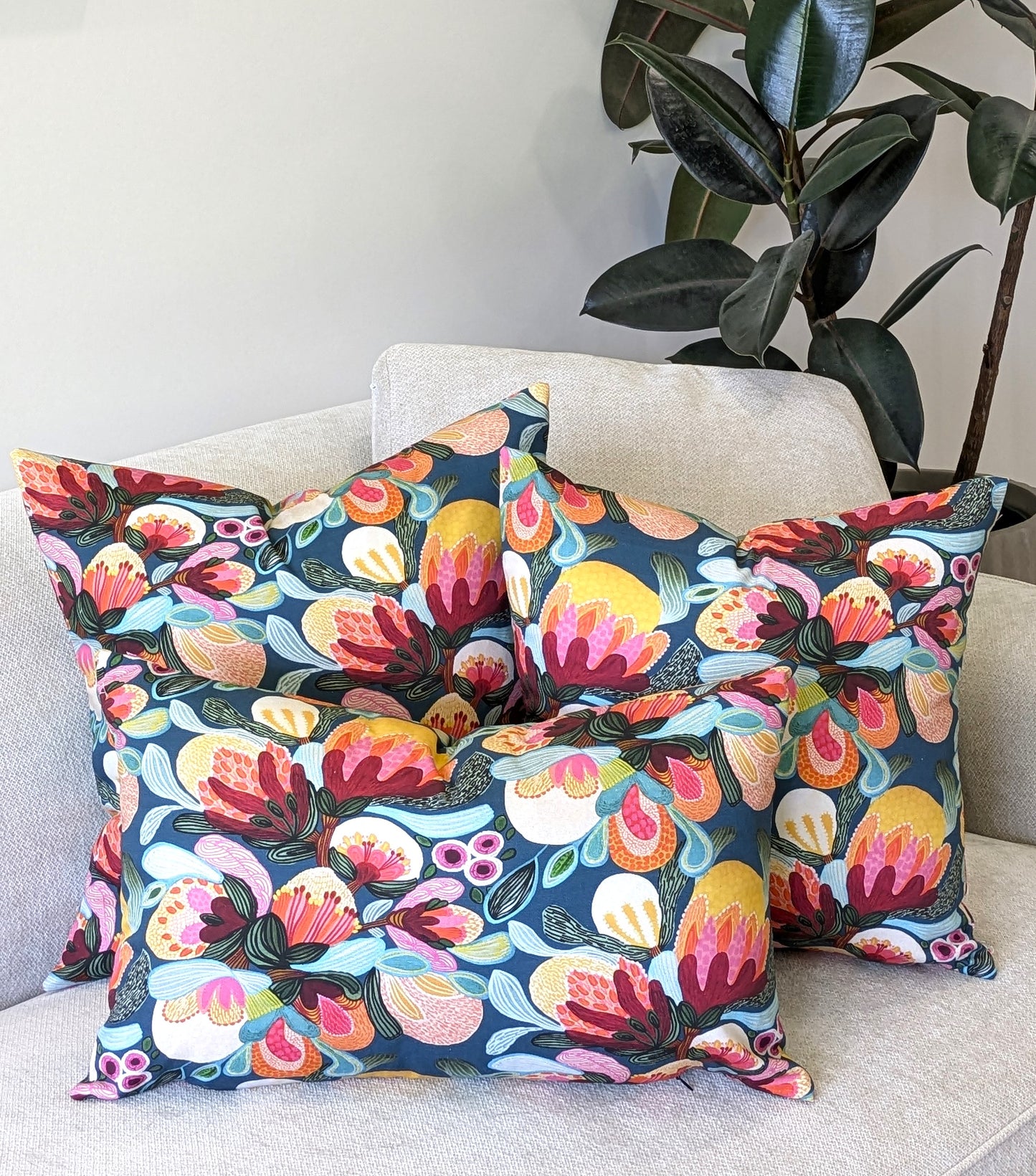 Three decorative pillows, including the Emotive Collections Midnight Bush Cushion Cover, feature bold floral patterns in pink, yellow, orange, and green on a light beige sofa with a leafy houseplant in the background.