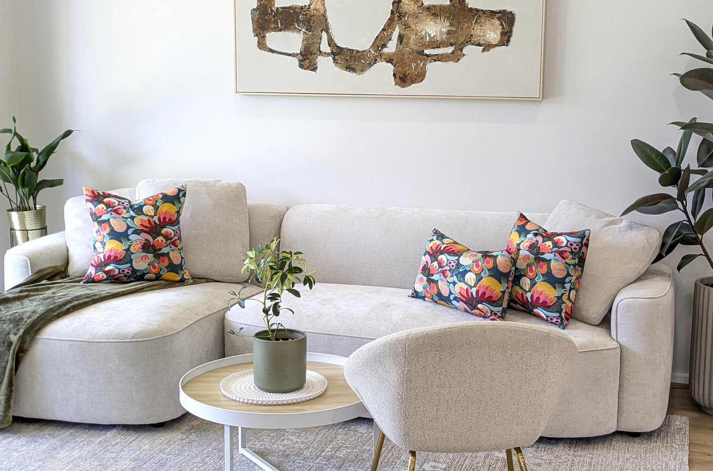 A modern living room with a light beige sectional, one Emotive Collection Midnight Bush Cushion Cover, a round ottoman with a potted plant, white armchair, green plants, and abstract wall art above the sofa.