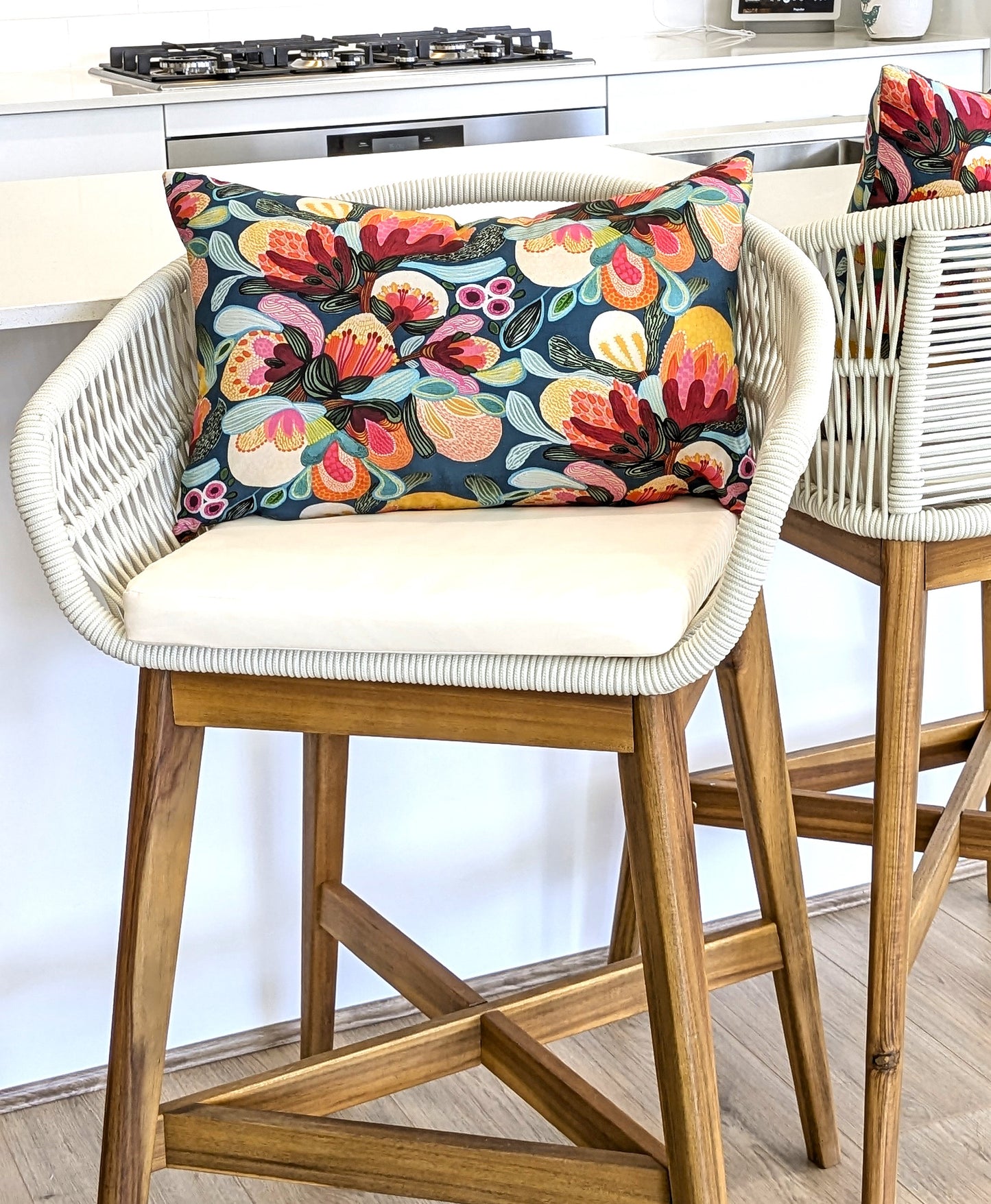 A white woven barstool with wooden legs is topped with Emotive Collection’s Midnight Bush Cushion Cover in cotton canvas, showcasing colorful florals. Another matching barstool sits nearby in a modern kitchen setting.