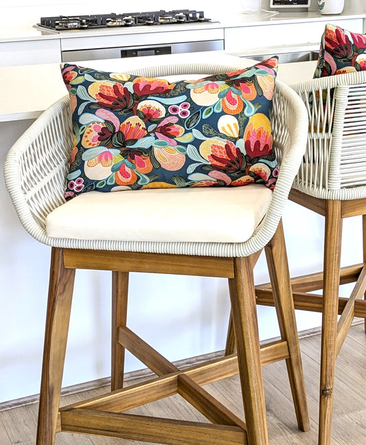 A white woven barstool with wooden legs is topped with Emotive Collection’s Midnight Bush Cushion Cover in cotton canvas, showcasing colorful florals. Another matching barstool sits nearby in a modern kitchen setting.