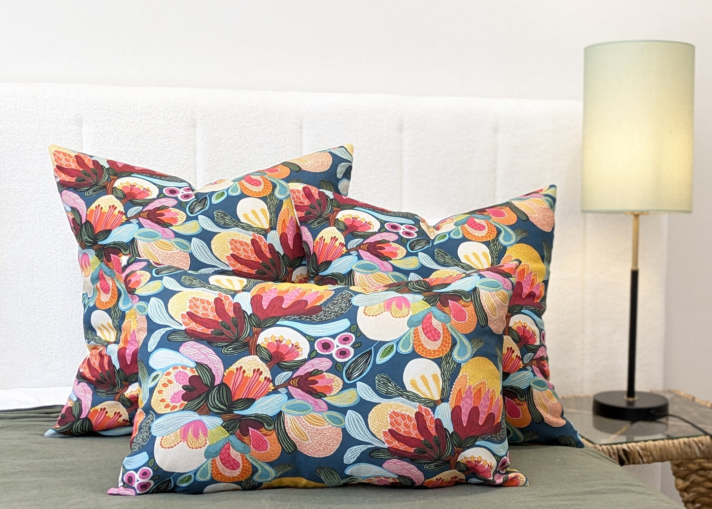 Three decorative pillows, including the Emotive Collections Midnight Bush Cushion Cover, are arranged on a bed with a white headboard. A lit light green lamp is on the bedside table to the right.
