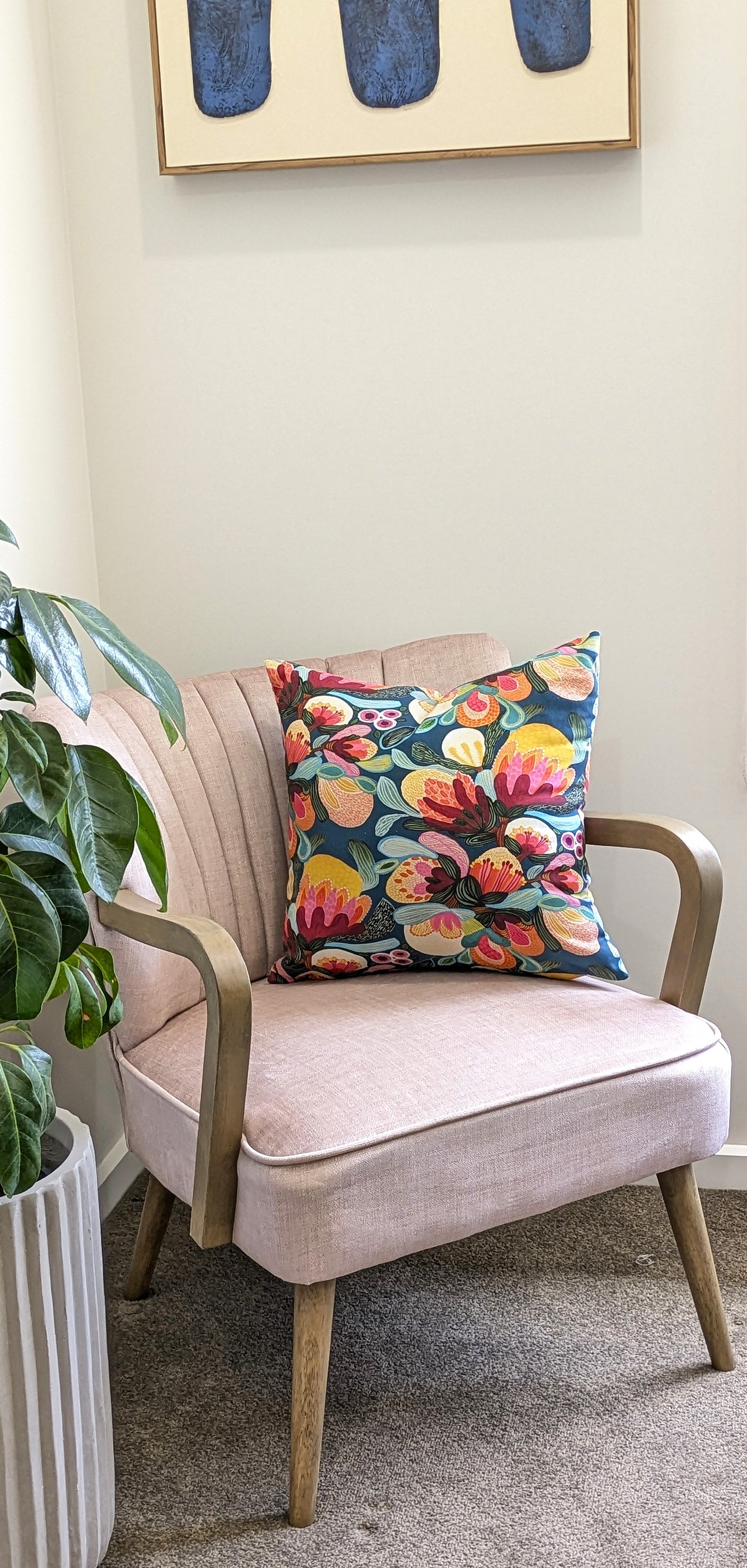 A light pink armchair with wooden arms and legs sits in a cozy corner. The Emotive Collection Midnight Bush Cushion Cover rests on the chair, complemented by a green potted plant and abstract art with blue shapes.