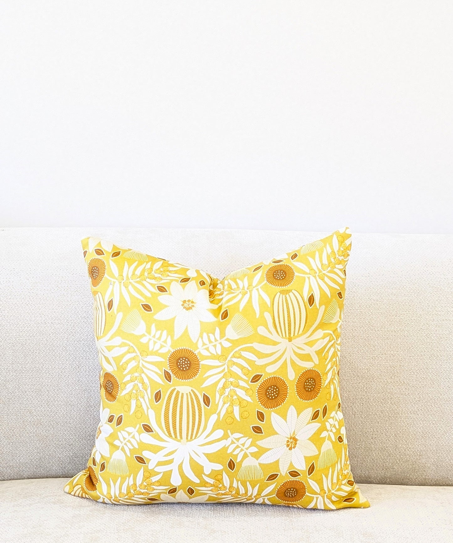 The Emotive Collection Wattle Mustard Cushion Cover, featuring a white and orange floral pattern, decorates a light sofa set against a plain white wall.
