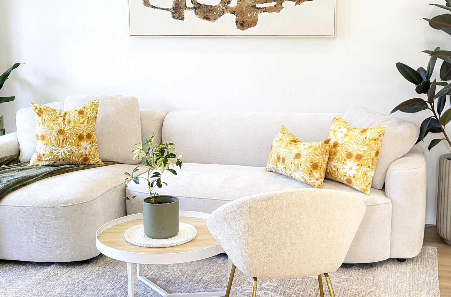 A modern living room with a cream sectional sofa, three Emotive Collection Wattle Mustard Cushion Covers, a round coffee table with a potted plant, a light armchair, lush greenery in large pots, and abstract wall art.