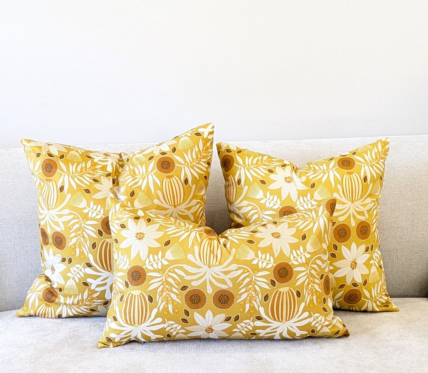Three yellow decorative pillows with white floral patterns, including the Emotive Collection Wattle Mustard Cushion Cover, are arranged on a light-colored couch against a plain white wall.