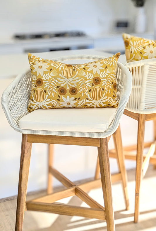 A white and wood barstool with a white cushion featuring the Emotive Collections Wattle Mustard Cushion Cover sits in a bright, modern kitchen. A matching stool with a handmade cover is partially visible beside it.