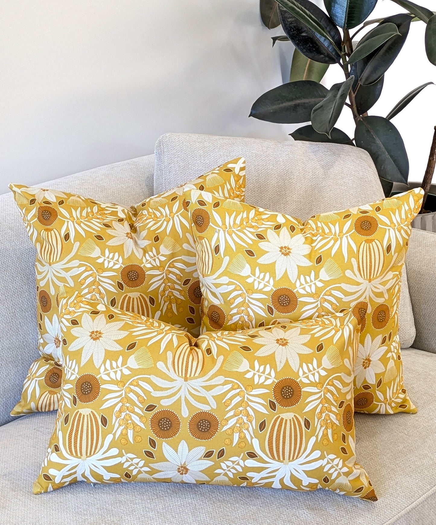 Three yellow decorative pillows, including the Emotive Collection’s Wattle Mustard Cushion Cover in white and brown floral design, are arranged on a light-colored sofa beside a green-leafed plant.