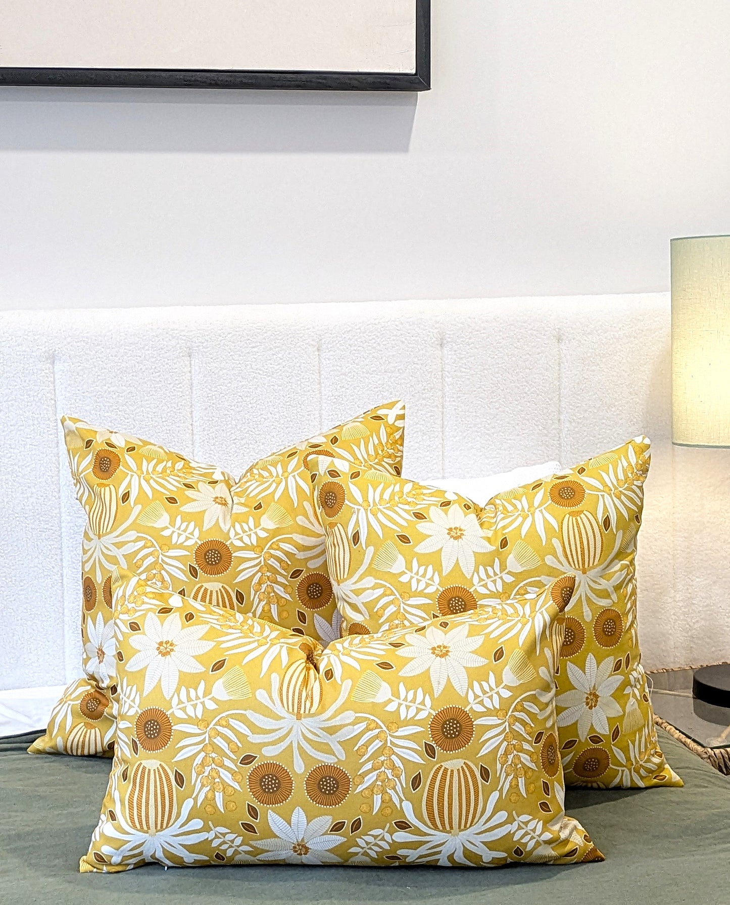 Three yellow and white floral throw pillows, including an Emotive Collection Wattle Mustard Cushion Cover, are arranged on a bed with a white upholstered headboard. A lamp sits on the bedside table and a framed picture hangs above.