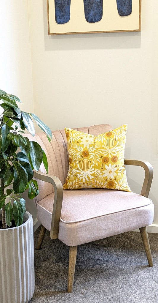 A pale pink upholstered armchair with wooden arms and legs sits in the corner, adorned with a Wattle Mustard Cushion Cover from Emotive Collection. Beside it is a large potted plant, and abstract wall art hangs above on the neutral wall.