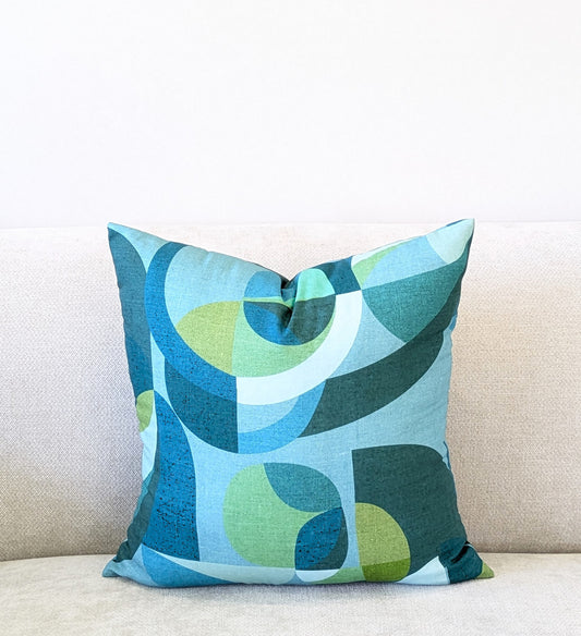 The Emotive Collection Linen Mid Century Blue Cushion Cover, featuring a modern geometric pattern in blue, teal, and green, rests on a light sofa against a plain white wall.