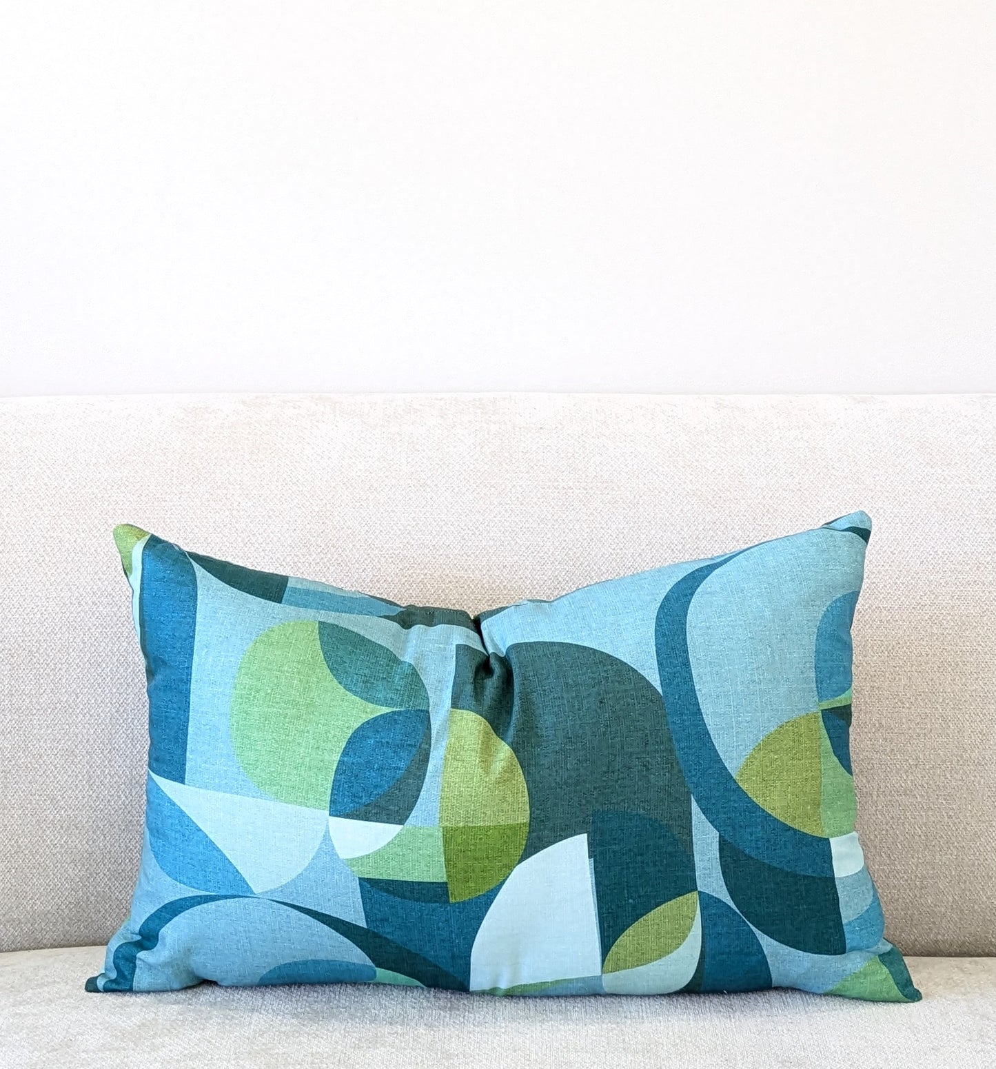 The Emotive Collection Linen Mid Century Blue Cushion Cover, featuring a blue and green geometric pattern, decorates a light beige sofa set against a plain white wall.