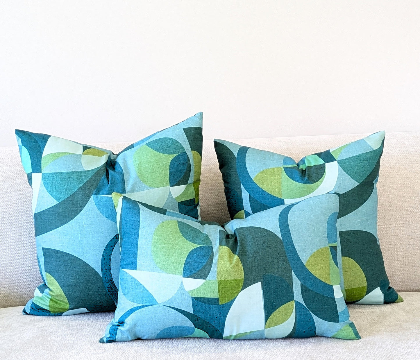Three Emotive Collection Linen Mid Century Blue Cushion Covers with blue, green, and white geometric patterns are displayed on a light couch against a plain white wall, each handmade from soft linen fabric.