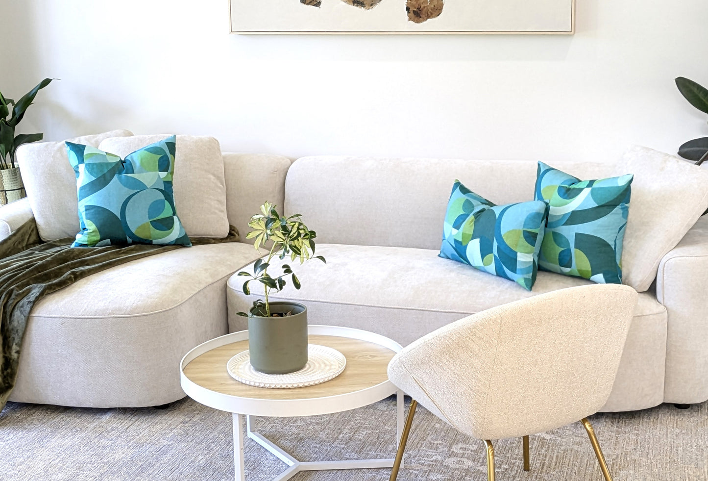 A modern living room with a light beige sofa featuring Emotive Collections Linen Mid Century Blue Cushion Cover, three blue and green abstract pillows, a round white coffee table with a potted plant, a cream chair, and lush green plants in the background.