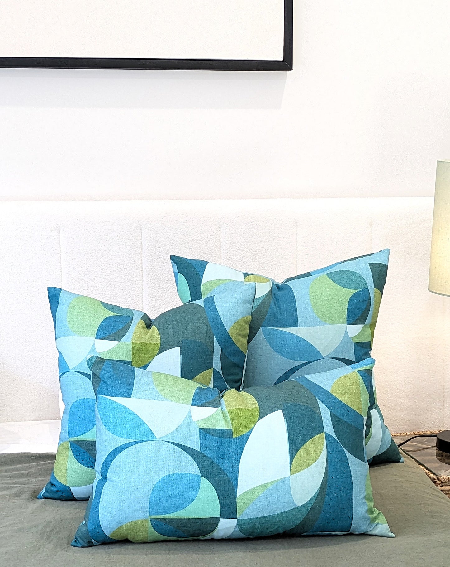 Four decorative pillows, including the Emotive Collection Linen Mid Century Blue Cushion Cover, display blue, green, and white abstract geometric patterns on a bed with a white headboard. A lamp and large frame are seen in the background.
