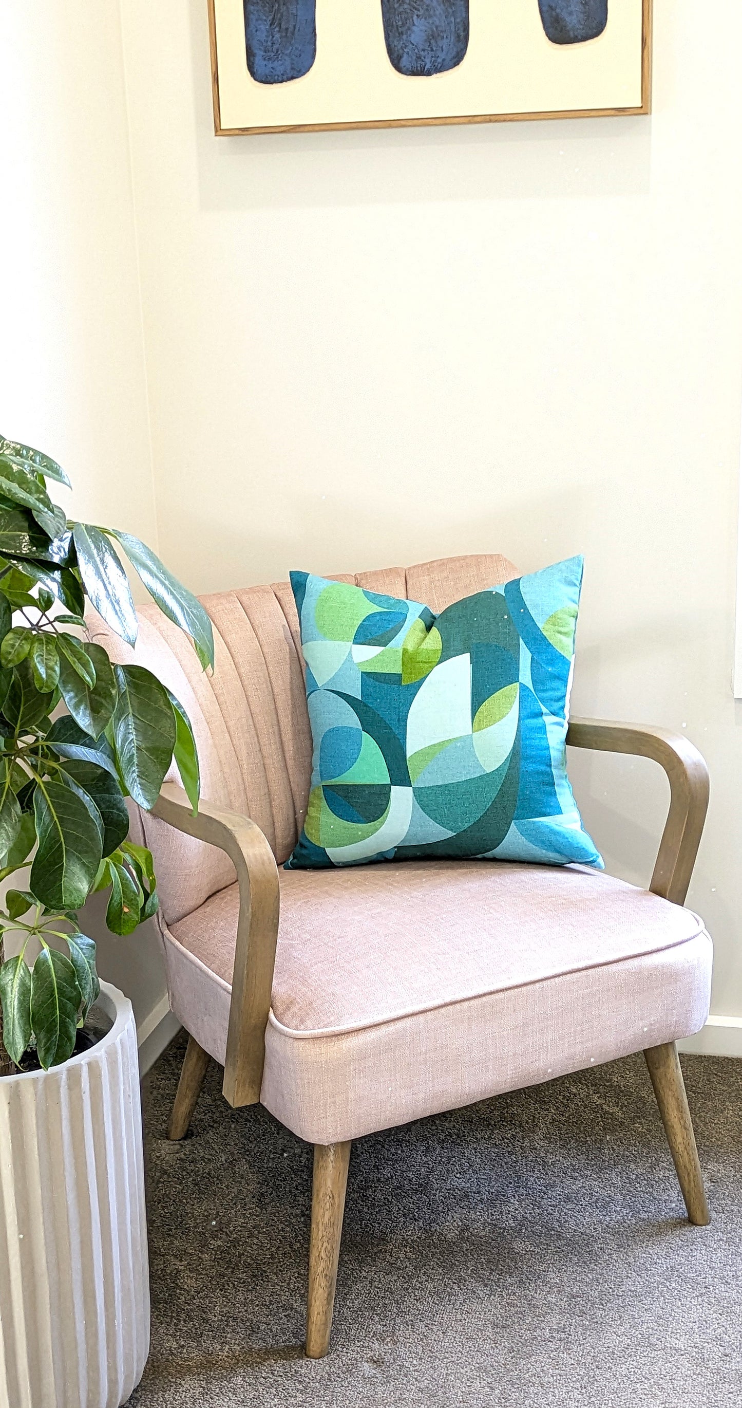 A light pink armchair with wooden legs sits beside a leafy potted plant. It features the Emotive Collections Linen Mid Century Blue Cushion Cover, handmade with colorful geometric patterns from soft linen. A modern painting hangs above.