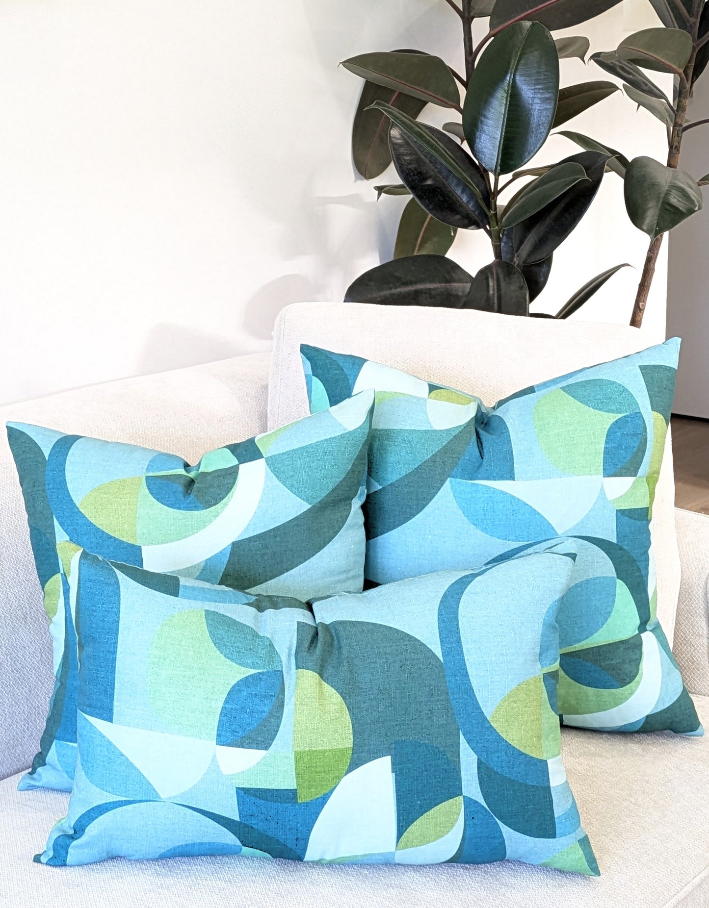 Four Emotive Collection Linen Mid Century Blue Cushion Covers, featuring blue, green, and teal mid-century designs, are arranged on a light sofa. A tall plant with dark green leaves completes this bright, modern setting.