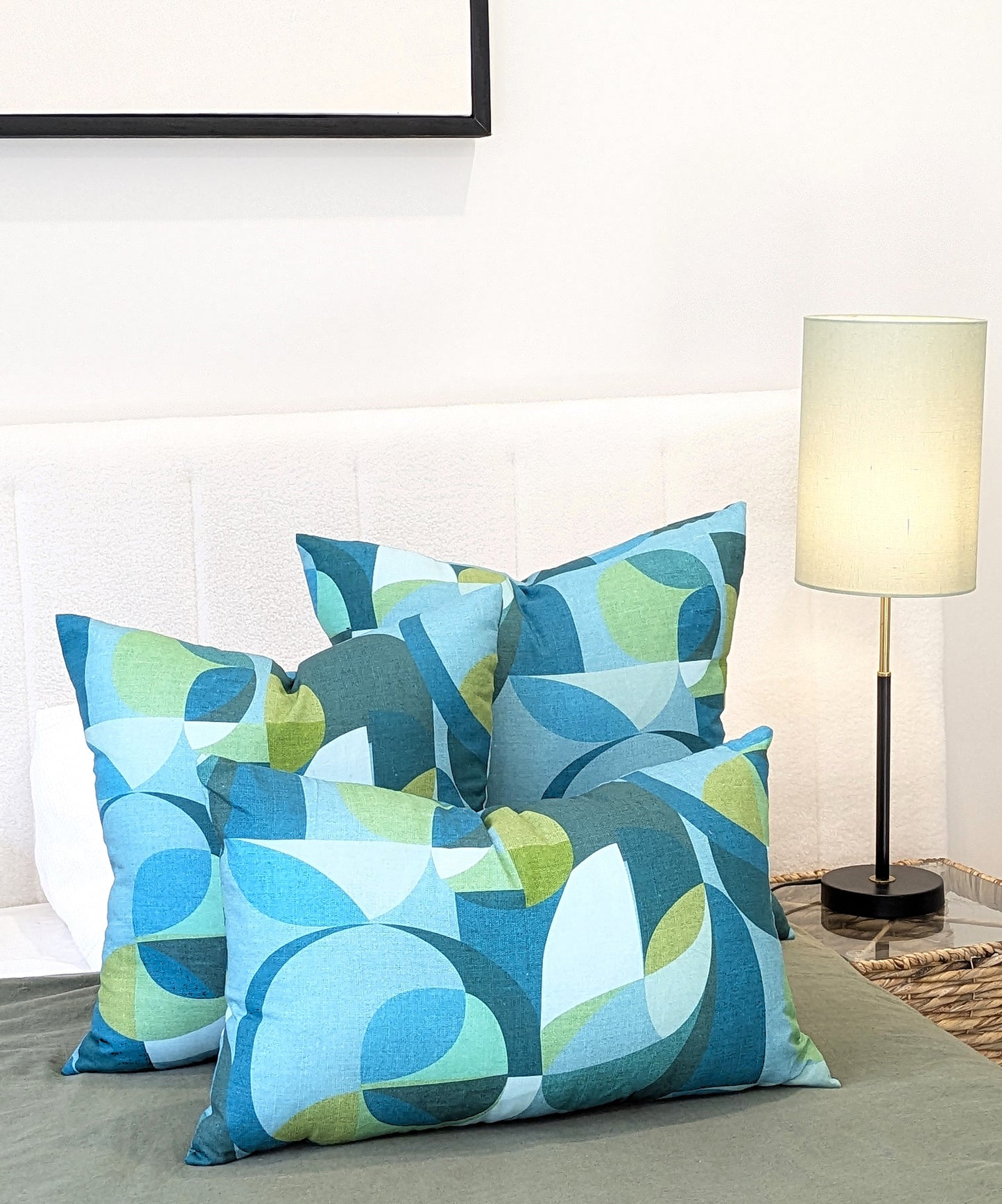 A neatly made bed showcases four geometric pillows, one featuring the Emotive Collection Linen Mid Century Blue Cushion Cover, beside a white headboard and a modern bedside lamp with a light green shade.