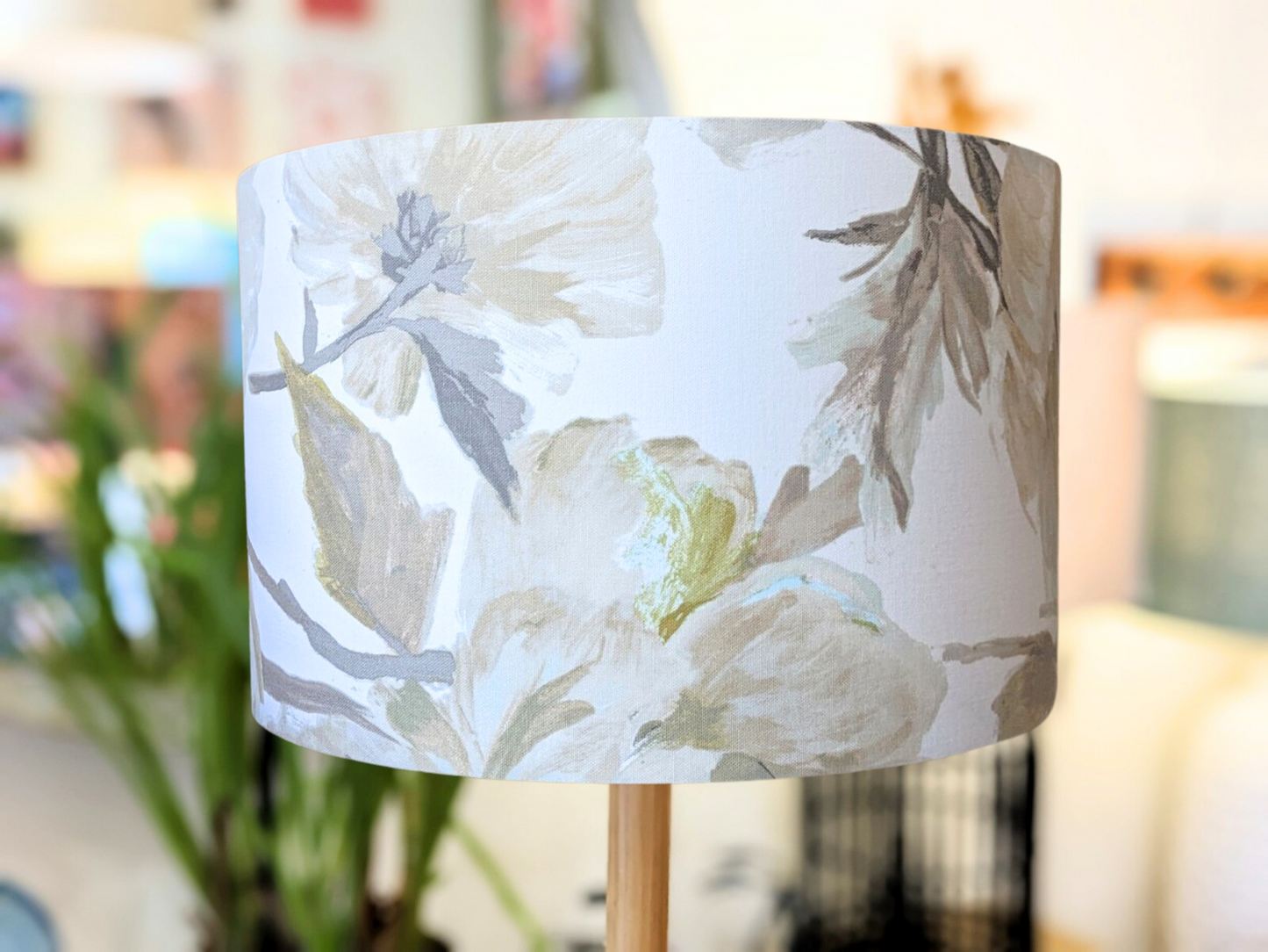 A close-up of the In Bloom Lampshade from the Emotive Collection features a soft floral pattern with large, muted flowers and leaves in beige, gray, and pale green—ideal for table lamps against a bright, blurred indoor backdrop.