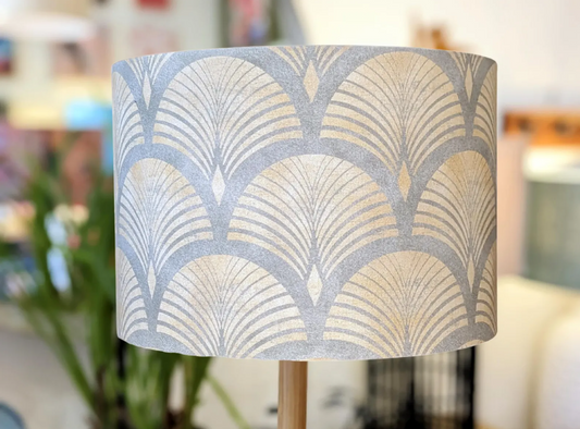 A close-up of the Emotive Collection Art Deco Lampshade, featuring a handcrafted geometric fan pattern in soft grey and beige tones. The background shows blurred indoor plants and furniture, captured in South Australia.