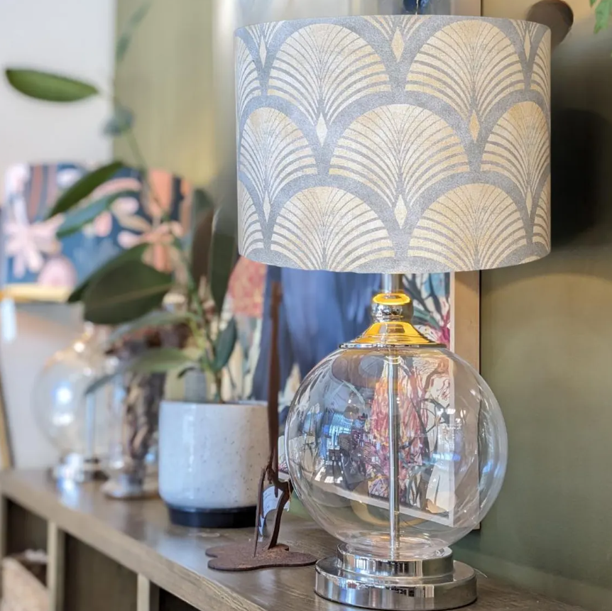 An Art Deco Lampshade from the Emotive Collection, featuring a modern clear glass base, sits on a wooden shelf surrounded by potted plants and decor, creating a cozy South Australia-inspired ambiance.