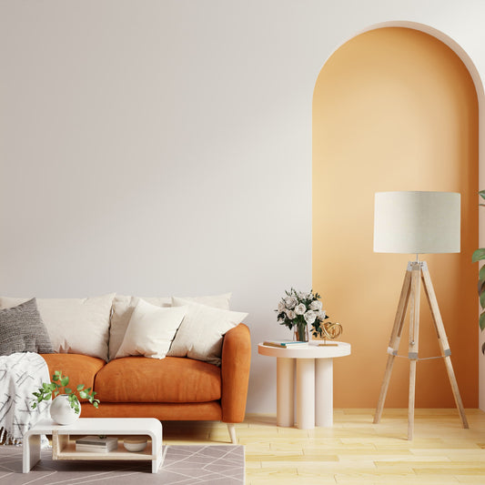 A modern living room with an orange sofa, beige and white cushions, and a round white coffee table. The Emotive Collection Cream Premium Linen Lampshade crowns a large tripod floor lamp, adding elegance against a white wall with a yellow arched accent.