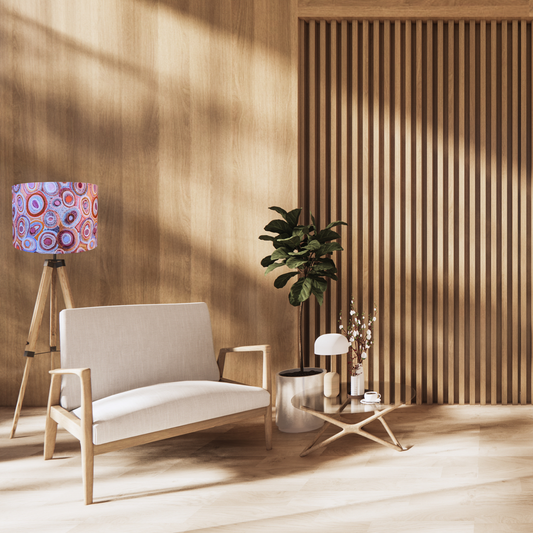 A modern, sunlit room with light wood walls and floor features a cream armchair, small table with decor, potted plant, and a tripod lamp topped with the Emotive Collection’s Water Dreaming Aboriginal lampshade.