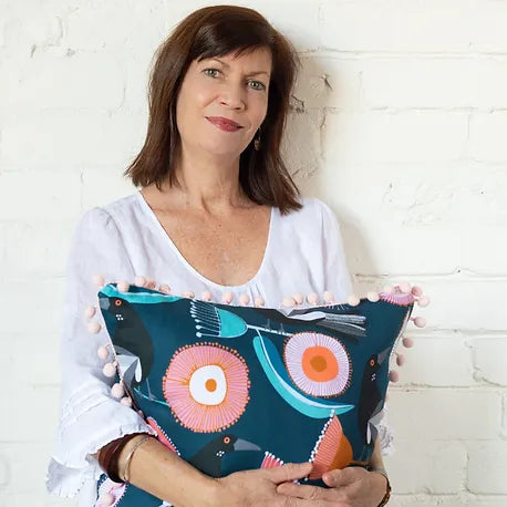 A woman with shoulder-length brown hair in a white blouse is smiling and holding a blue cushion with colorful, abstract floral patterns and pom-pom trim, standing against a white brick wall.