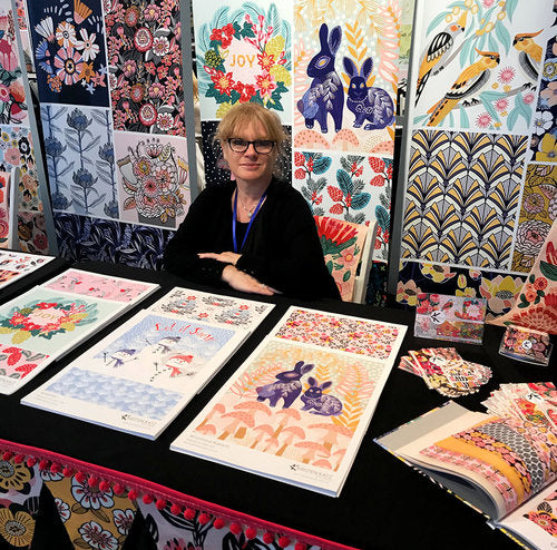 A woman with glasses and short blonde hair sits at a table displaying colorful art prints and cards with floral, animal, and nature designs. More vibrant artwork is displayed on the wall behind her.