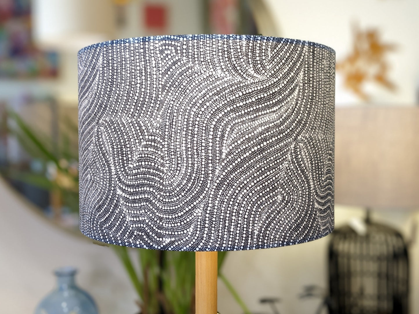 A close-up of the Desert Dreaming Aboriginal lampshade by Artist Senita Napangardi Granites, handmade with a wavy black and white dotted pattern, shown indoors against a softly blurred background with home decor and a mirror.
