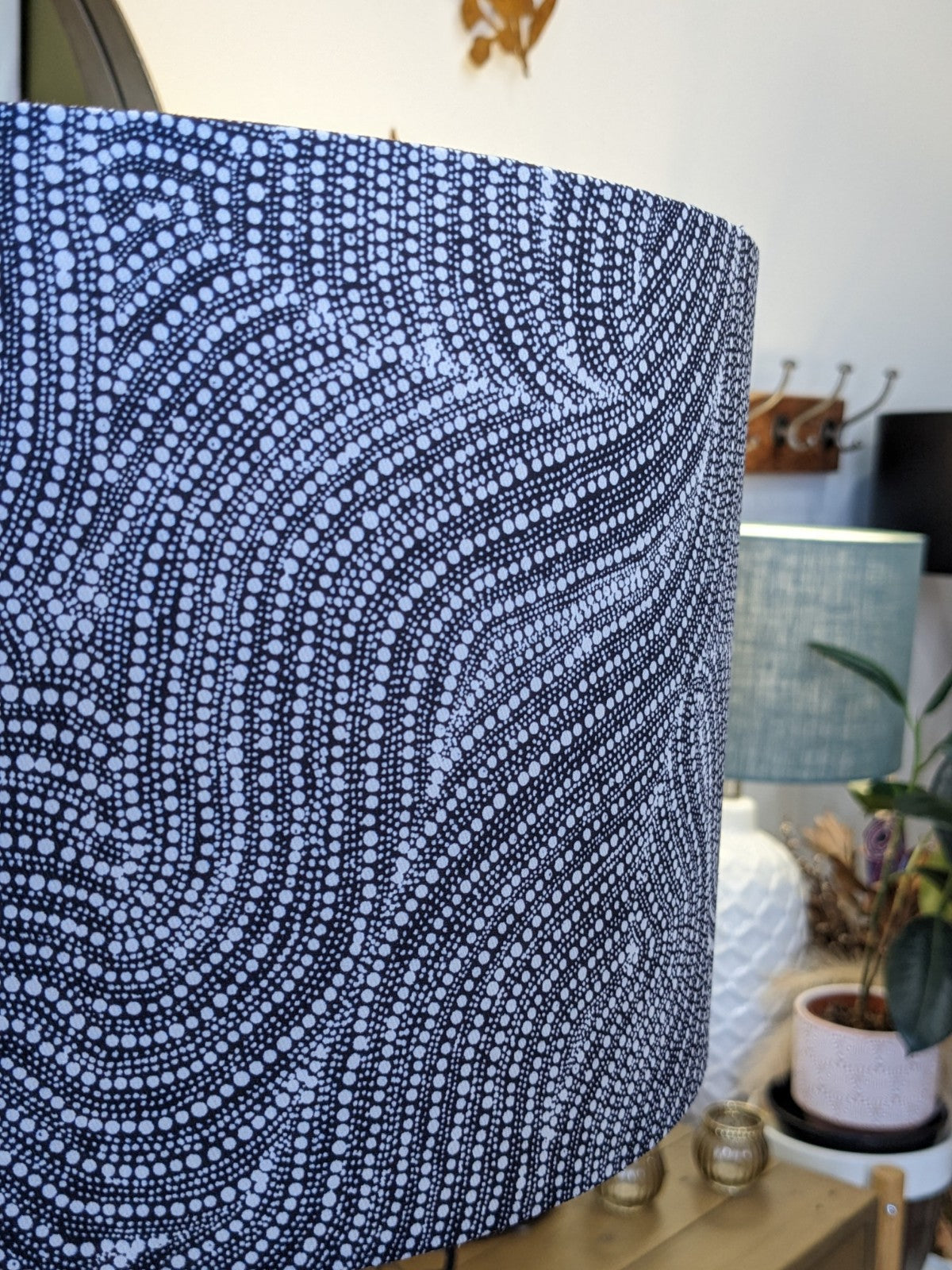 A close-up of the Desert Dreaming Aboriginal lampshade by Artist Senita Napangardi Granites, featuring a black and white dotted wavy pattern. Other lamps, a plant, coat rack, and mirror appear in the softly lit room.
