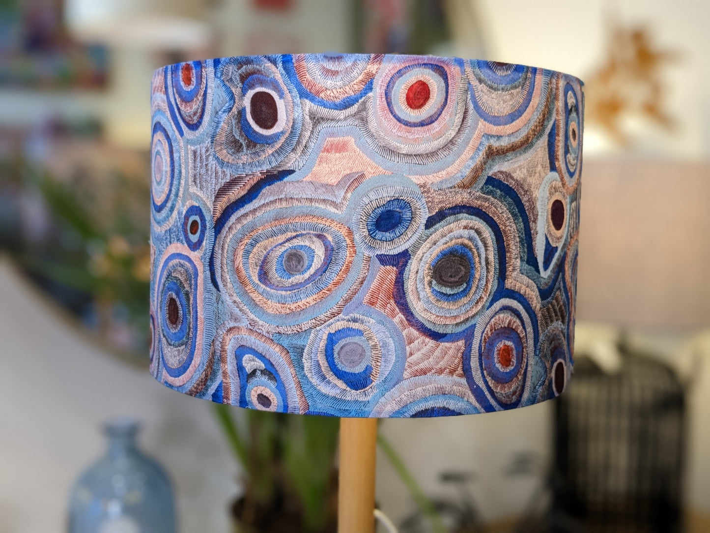The Aboriginal Water Dreaming Pastel Lampshade by artist Senita Napangardi Granites features abstract, circular patterns in blue, gray, and red with concentric rings and textured embroidery on a softly blurred background.