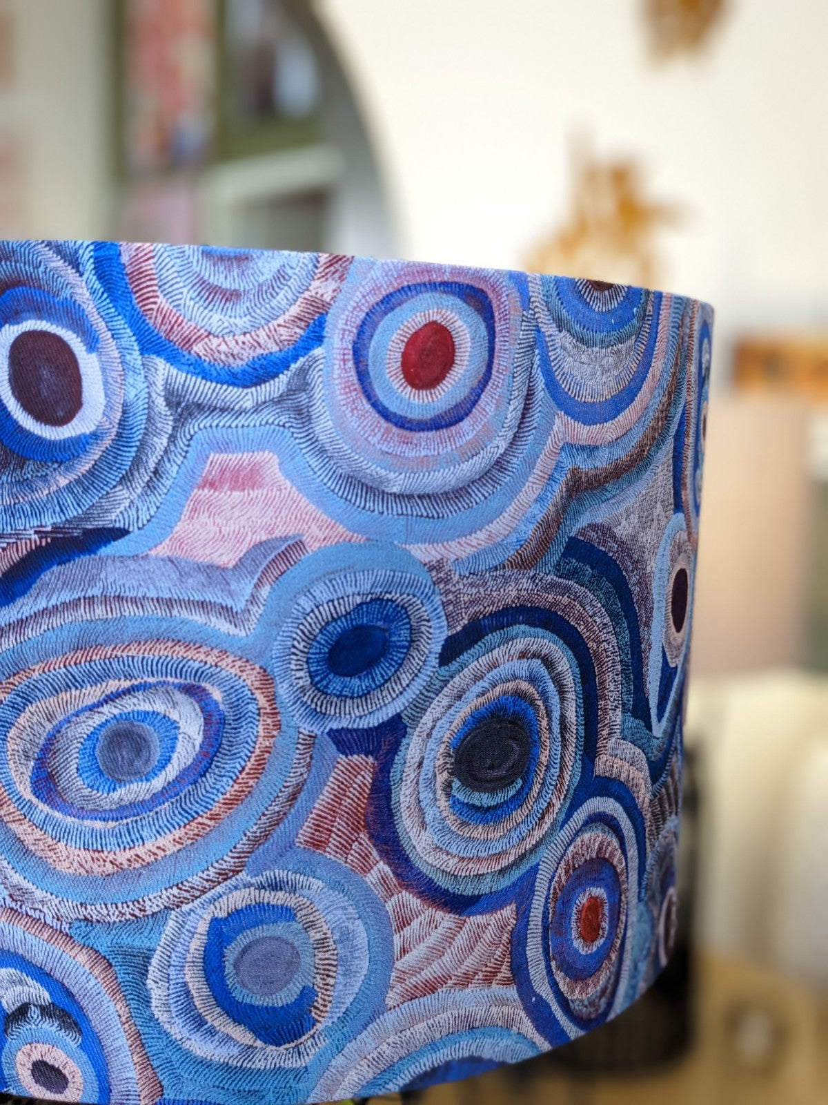 A close-up of the Aboriginal Water Dreaming Pastel Lampshade by artist Senita Napangardi Granites, featuring concentric circular patterns in blue, gray, and pink against a blurred interior background.