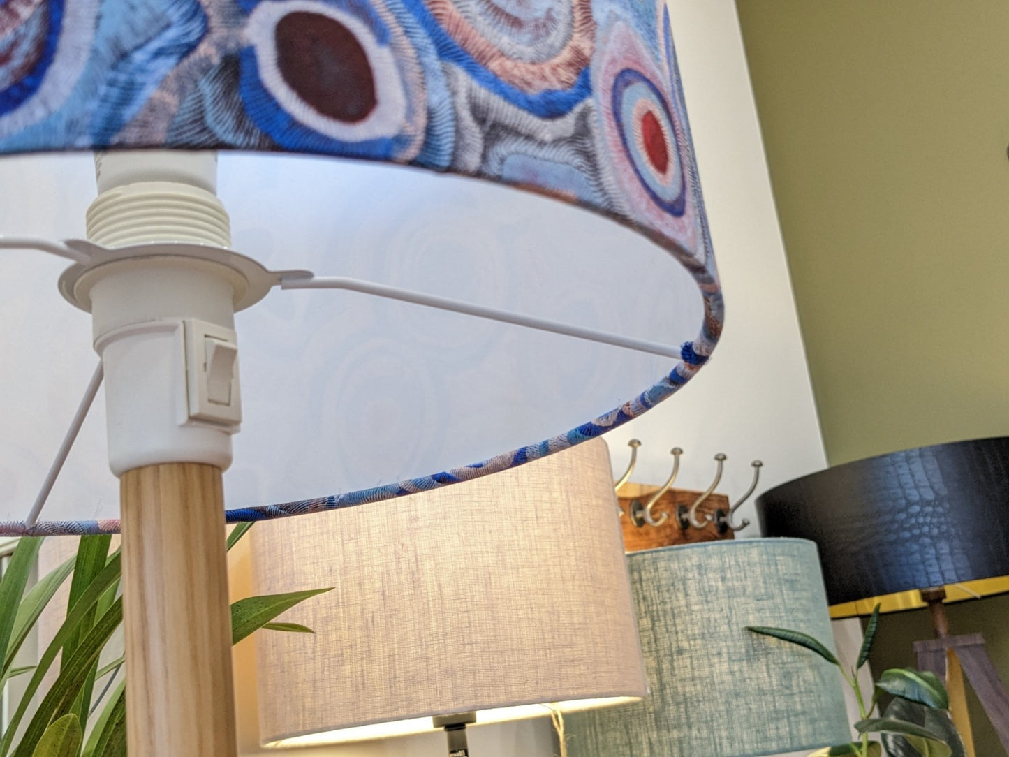 A close-up of the Aboriginal Water Dreaming Pastel Lampshade by artist Senita Napangardi Granites. In the background, lamps with beige, green, and black shades sit among green plants, inspired by Desert Fringe-Rush Seed Dreaming.