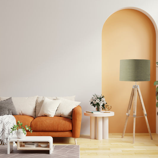 A modern living room with an orange sofa, neutral cushions, a white coffee table, and a tall floor lamp topped with the Emotive Collections Sage Premium Linen Lampshade. An arched soft yellow accent wall adds extra warmth.