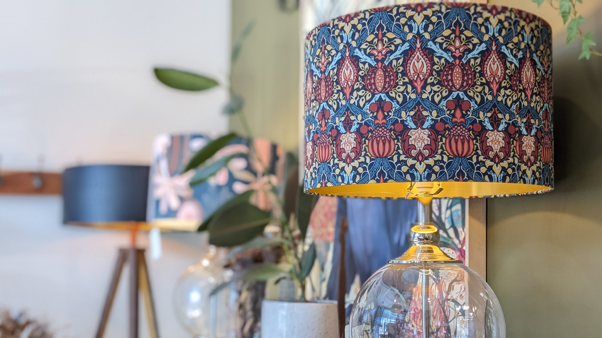 A clear glass table lamp with a colorful, floral-patterned lampshade sits in the foreground; two more lamps with similar shades are blurred in the background. Green leaves add a touch of natural decor.