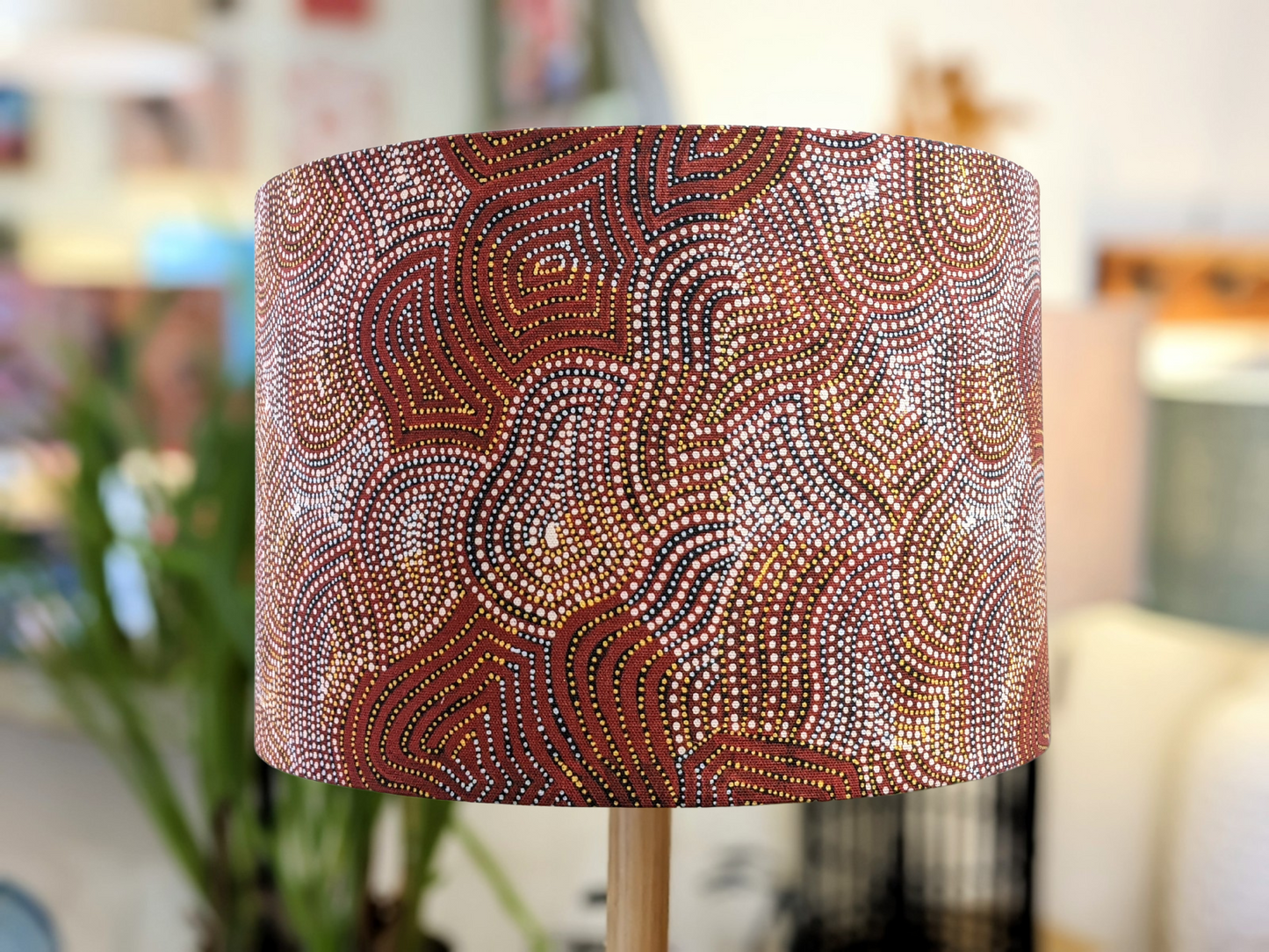 The “Warlu Mina Mina Jukurrpa (Ngalyipi)” lampshade features Mekeisha Napanangka Martin’s intricate Indigenous Aboriginal design in earthy brown, white, red, and yellow tones, set against an indoor backdrop of furniture and plants.