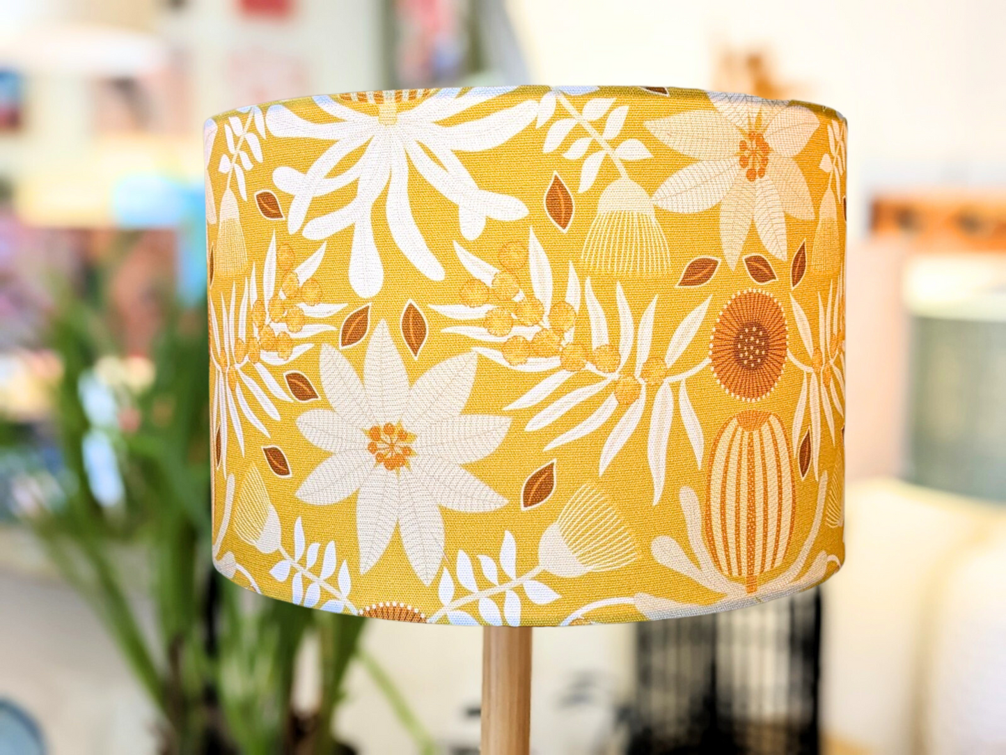 A close-up of the Emotive Collection’s Native Flora Mustard Lampshade, featuring white and brown floral and leaf patterns on a yellow background by Jocelyn Proust, set indoors with a softly blurred backdrop.
